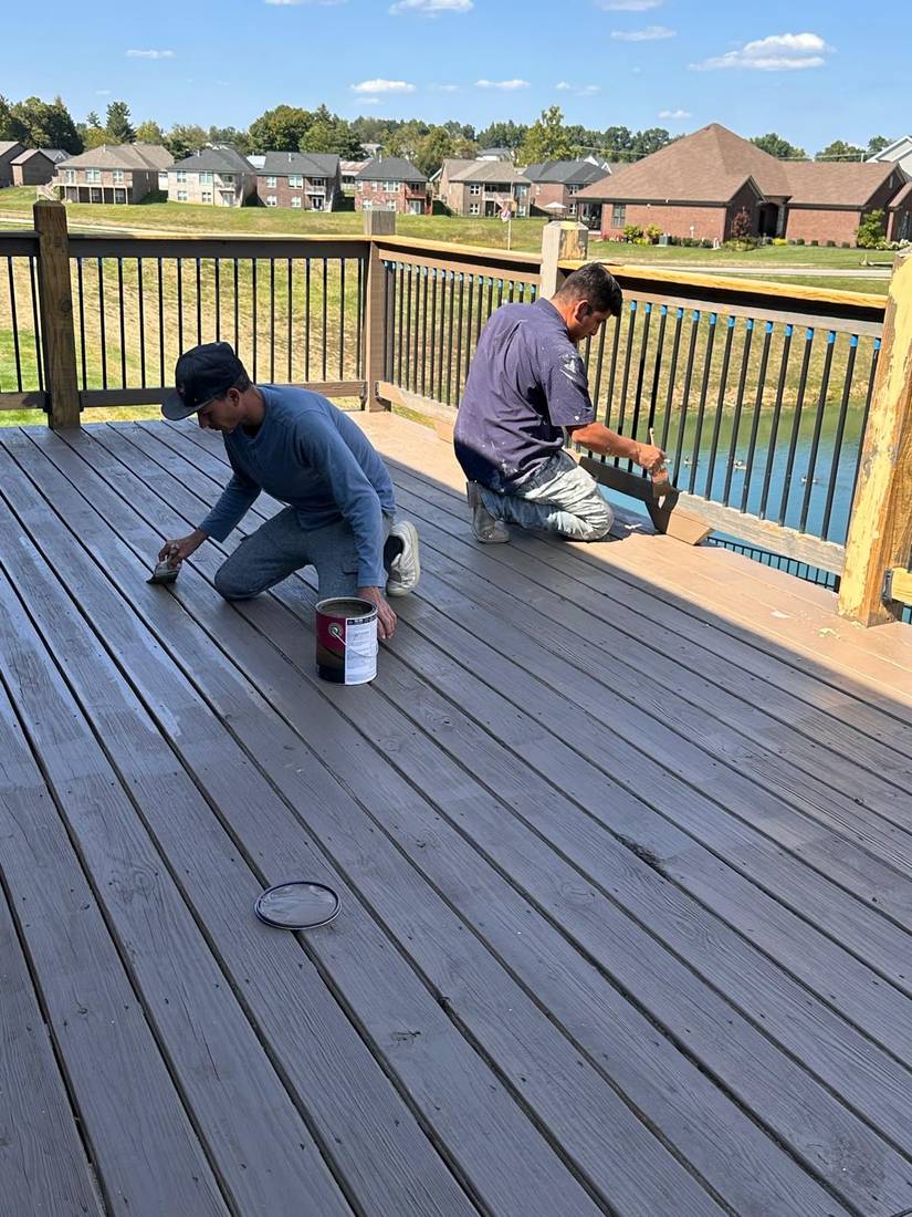 Two men are kneeling on a wooden deck, applying stain with brushes while overlooking a lake.