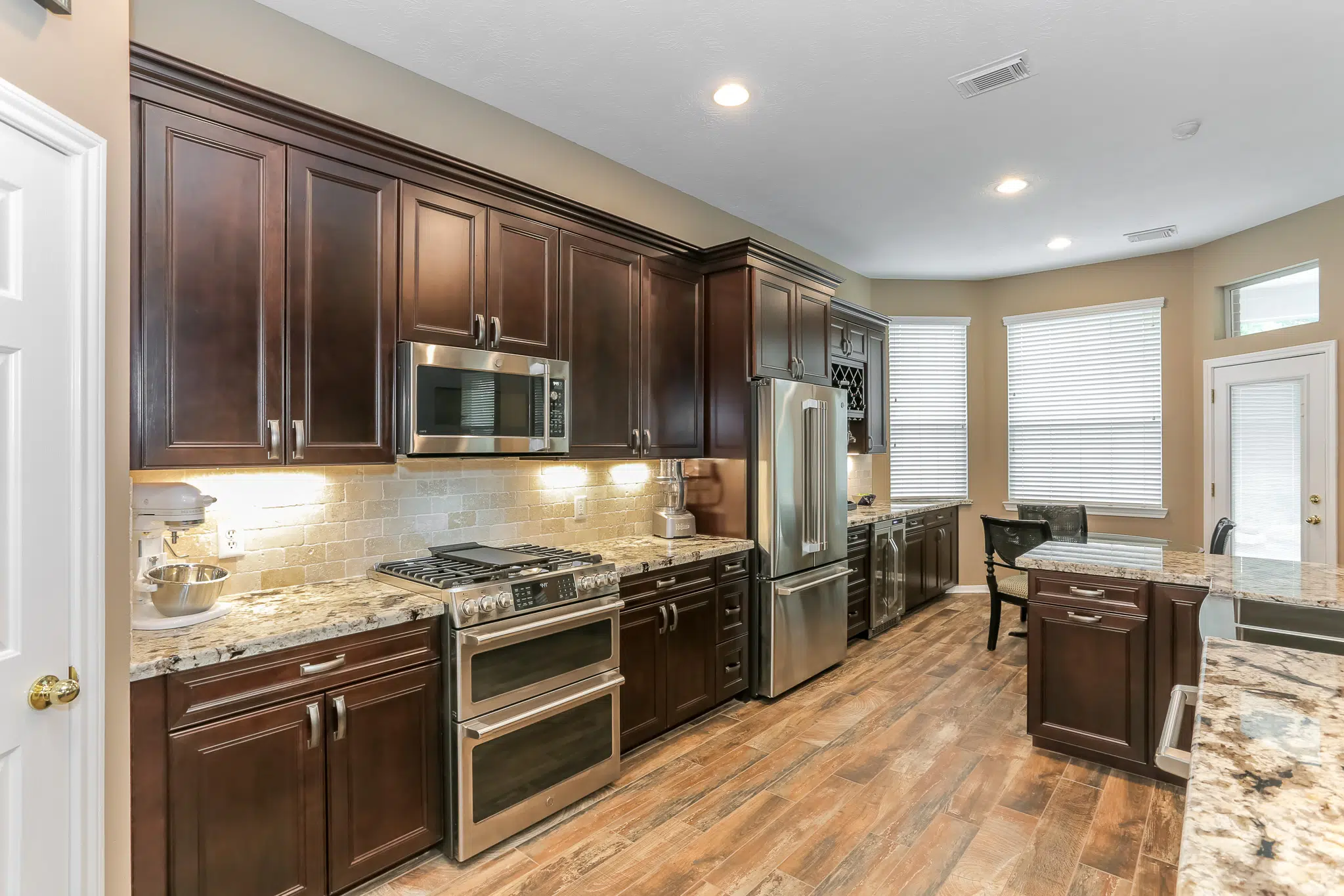 A spacious kitchen featuring dark wood cabinets, stainless steel appliances, and granite countertops under warm lighting.